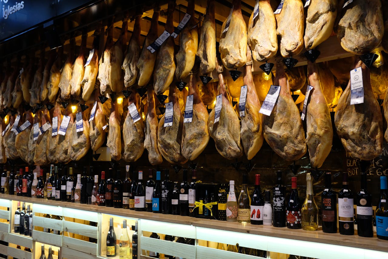 A vibrant display of hanging Spanish jamón with wine bottles in a Barcelona market.